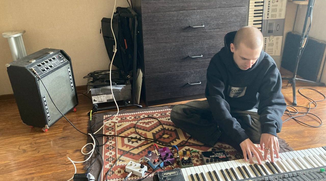 Музыкант сидит на полу с синтезаторной клавиатурой, рядом педали и комбоусилитель